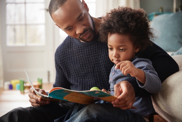 A Black father and his young son read a book together at home. 