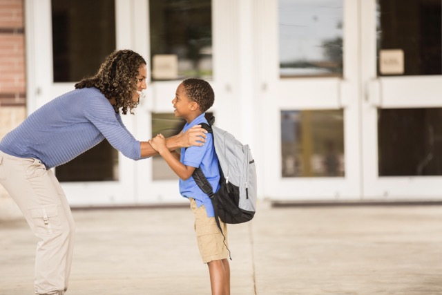 A Black elementary school student mom's arm for reassurance as she touches his shoulder and smiles in front of a school.