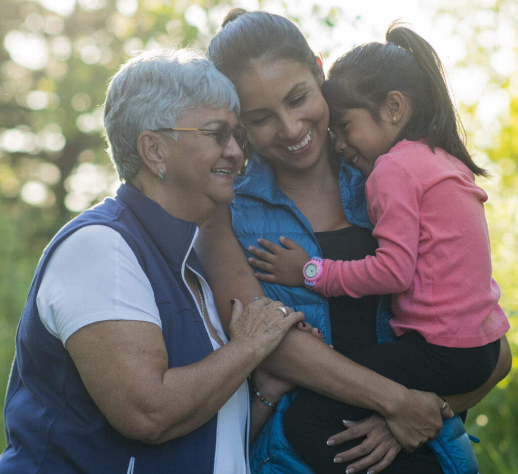 A young Hispanic ethnicity family go hiking in the forest at sunset. The toddler, her mother, and grandmother are all hugging and smiling at each other in active wear.
